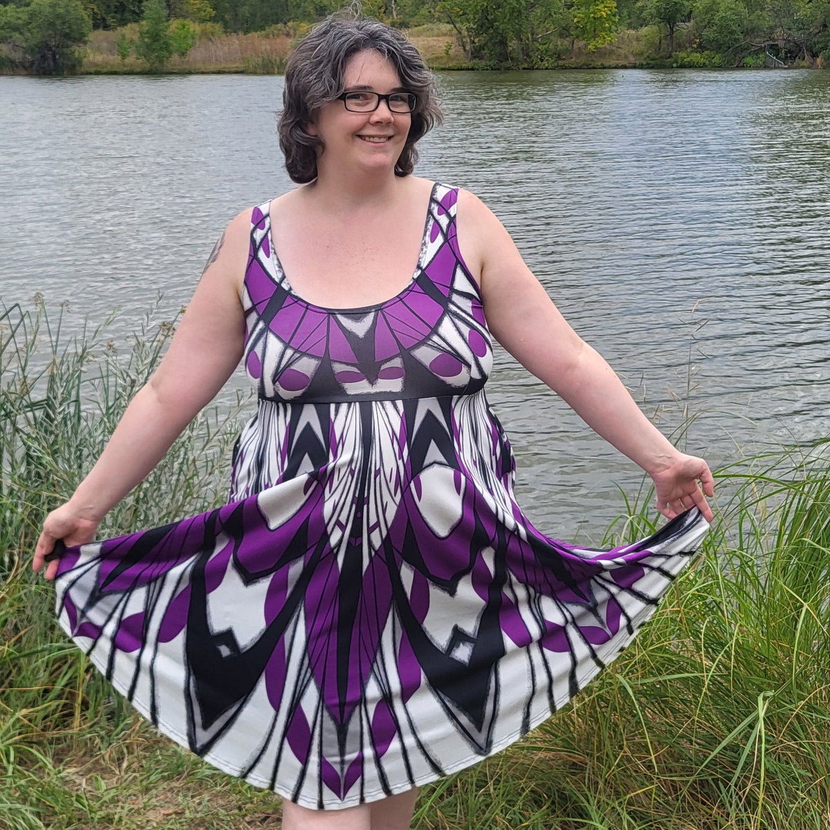 A woman wearing a sleeveless purple, black, and white patterned dress stands outdoors by a lake, holding out her skirt to show its full flow. Trees and a cloudy sky are visible in the background.