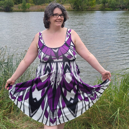 A woman wearing a sleeveless purple, black, and white patterned dress stands outdoors by a lake, holding out her skirt to show its full flow. Trees and a cloudy sky are visible in the background.